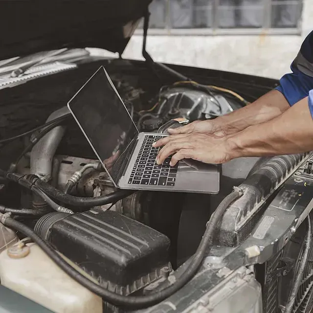 Un hombre está usando una computadora portátil debajo del capó de un automóvil.