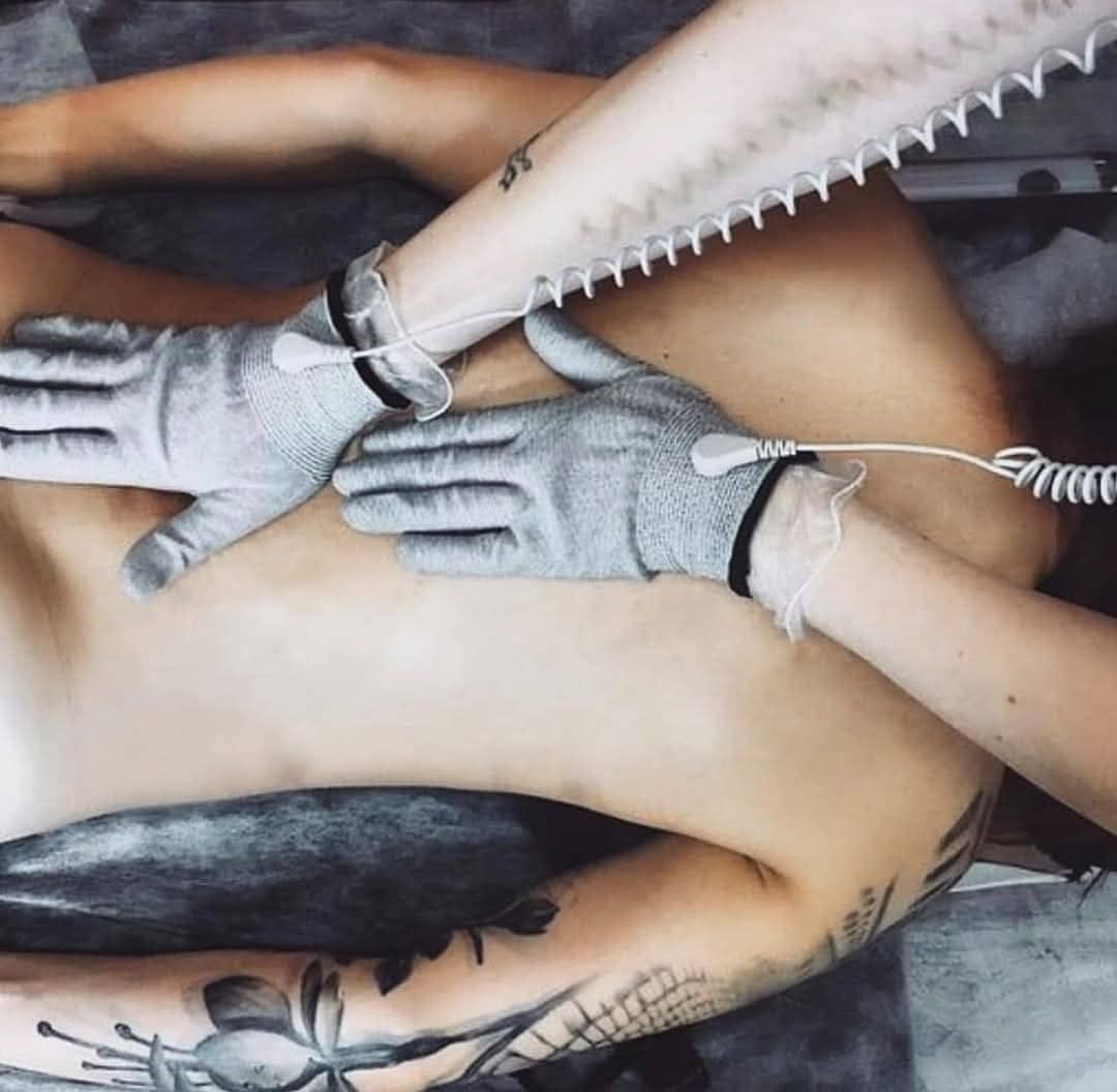 Manos enguantadas presionando la espalda desnuda de una persona durante una sesión de tatuaje, con los brazos tatuados visibles arriba y abajo.