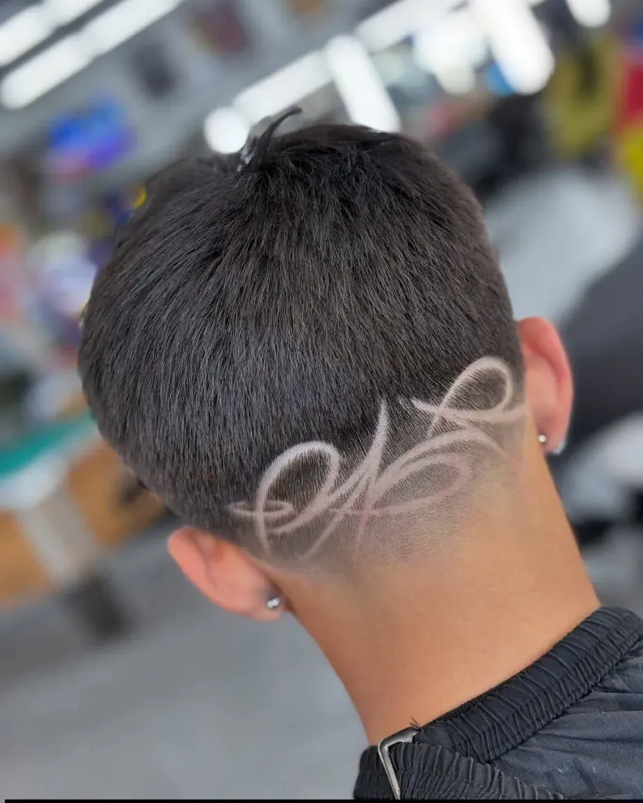 Hombre cortándose el pelo; primer plano de un degradado y una raya al medio. La mano del barbero señala la oreja, en el interior de una barbería.