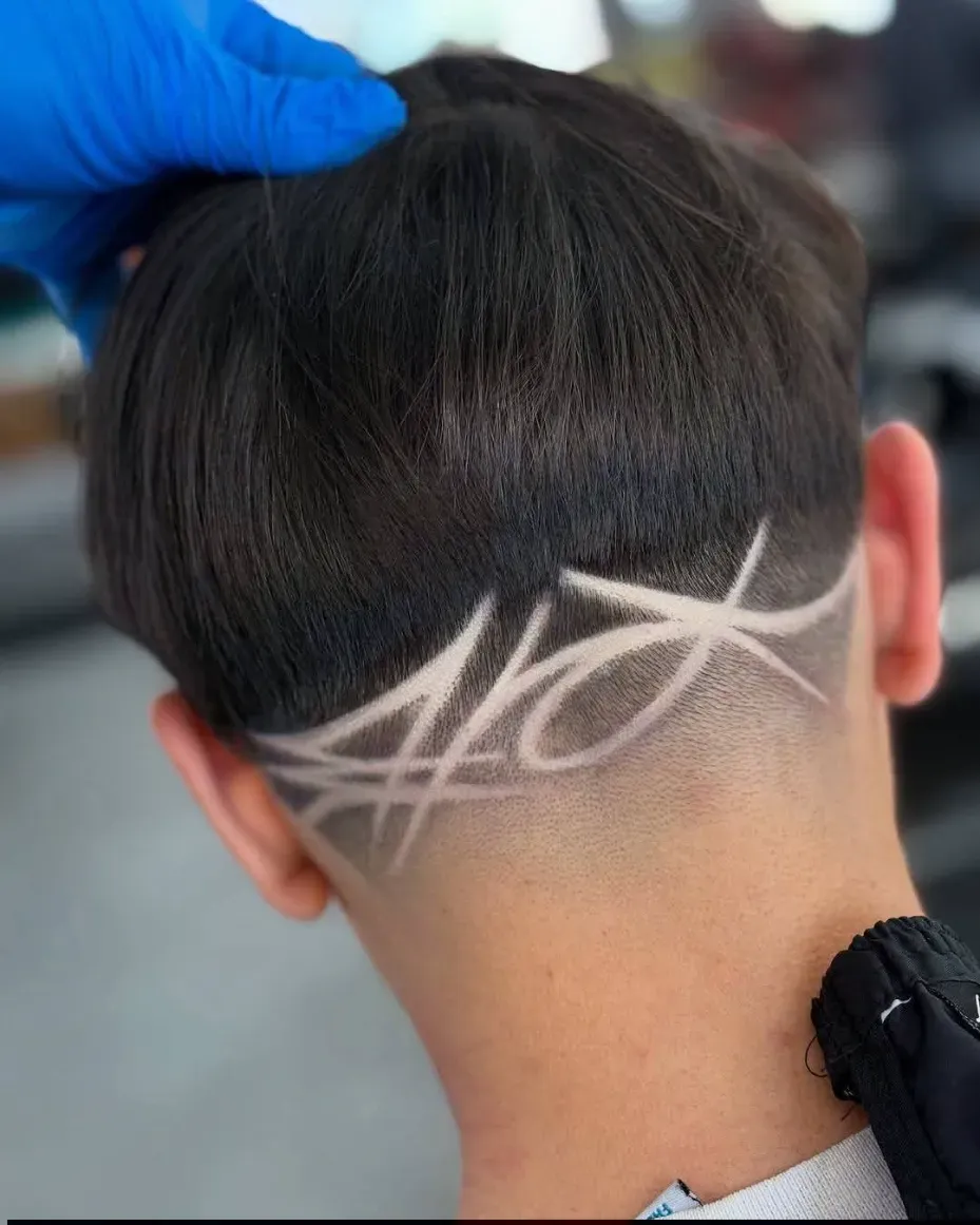 Primer plano de la nuca de una persona con un diseño afeitado en su cabello oscuro; una mano enguantada toca su cabeza.