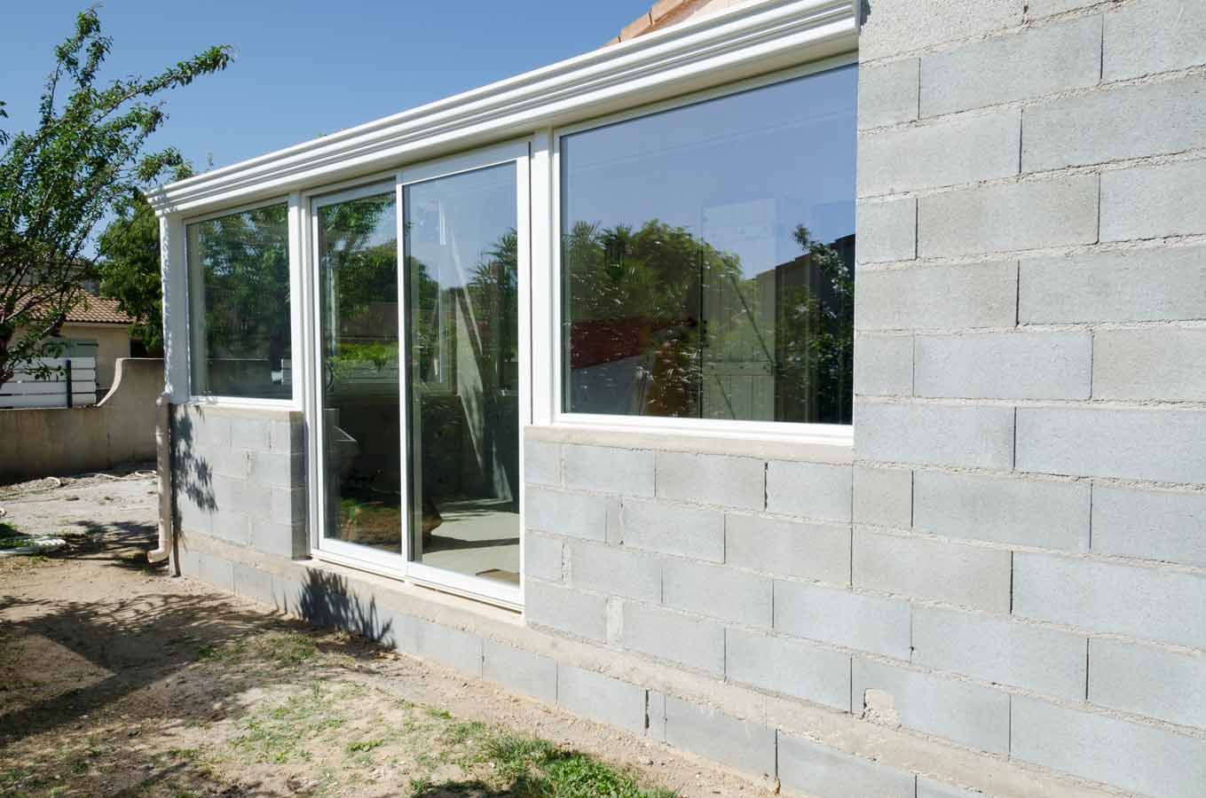 Maison en parpaings partiellement finie, avec de grandes portes coulissantes en verre et des boiseries blanches.