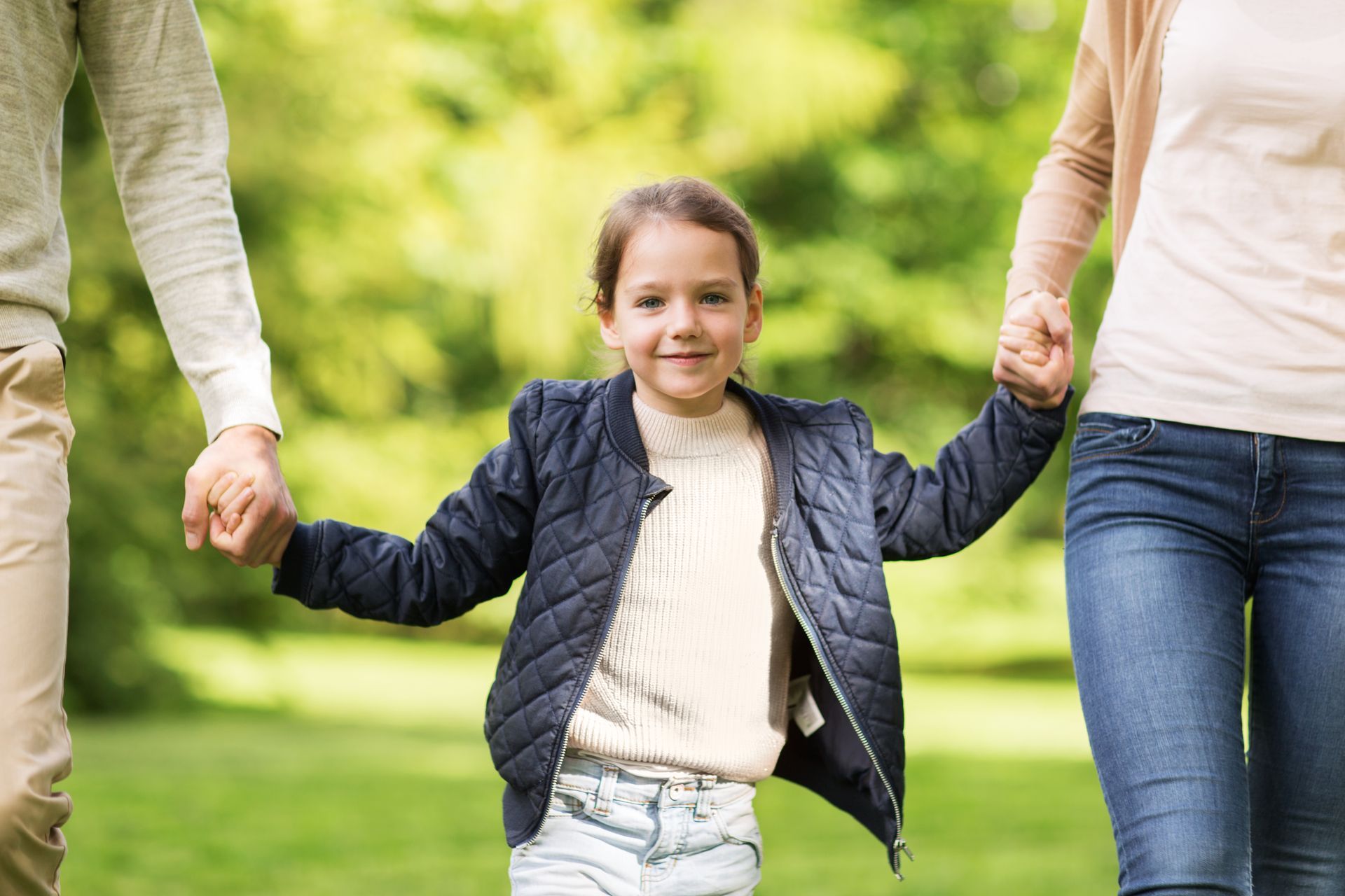 Un enfant souriant tenant la main de deux adultes dans un parc verdoyant.