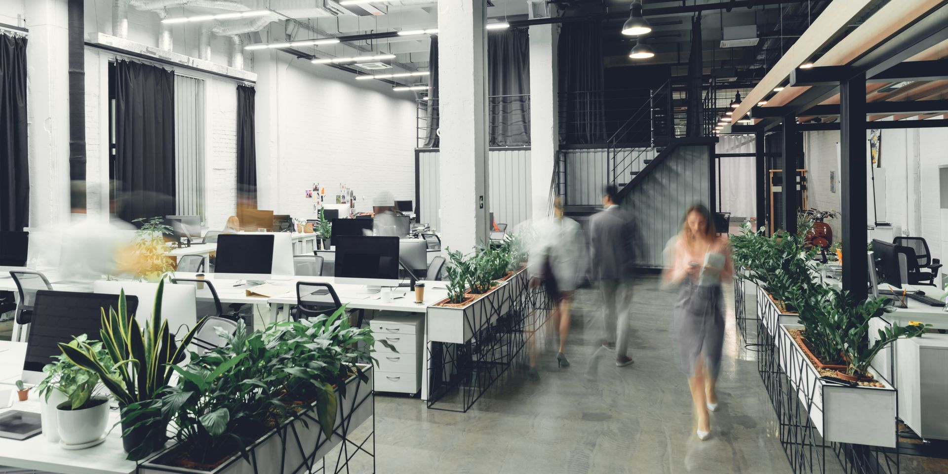 Bureau moderne à aire ouverte avec bureaux, plantes et deux personnes floues qui traversent l'allée.