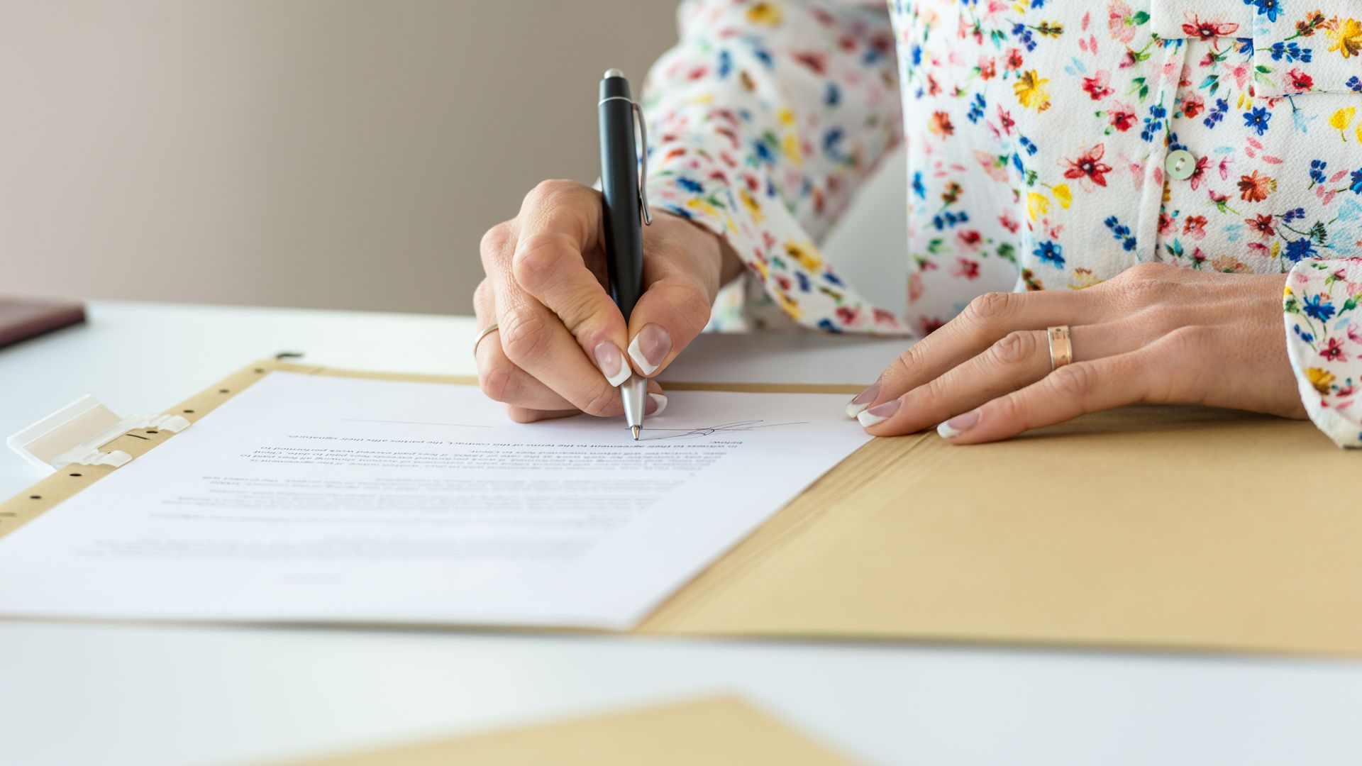 Une personne signe un document au stylo, assise à un bureau, vêtue d'une chemise à fleurs.