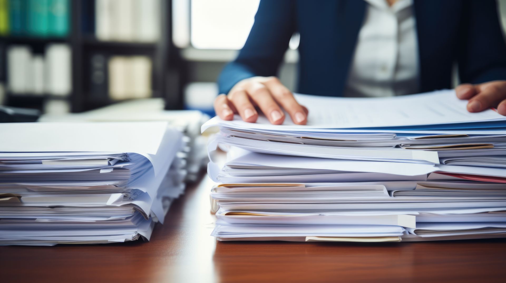 Des piles de documents papier sur un bureau, une personne triant des dossiers en arrière-plan.