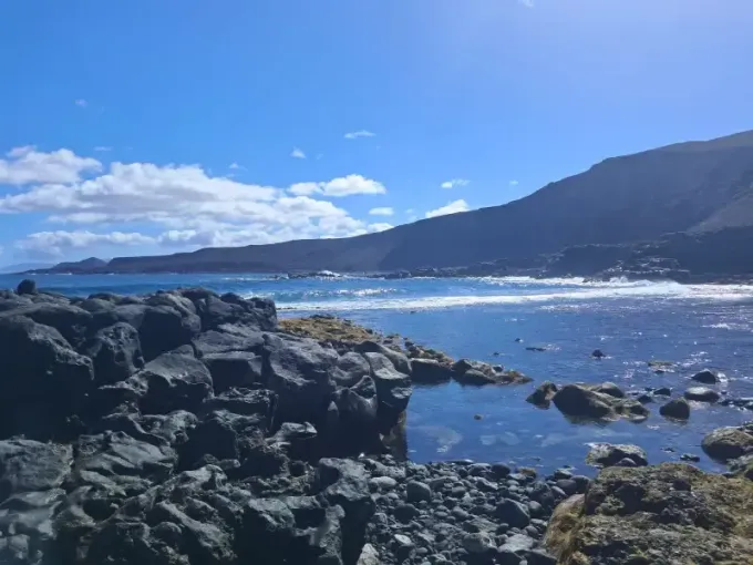 Una costa volcánica rocosa y oscura que se funde con el océano azul brillante bajo un cielo despejado con nubes blancas