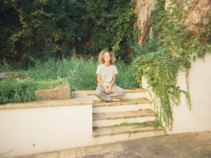 Una persona con cabello rizado está sentada con las piernas cruzadas en unos escalones de piedra al aire libre