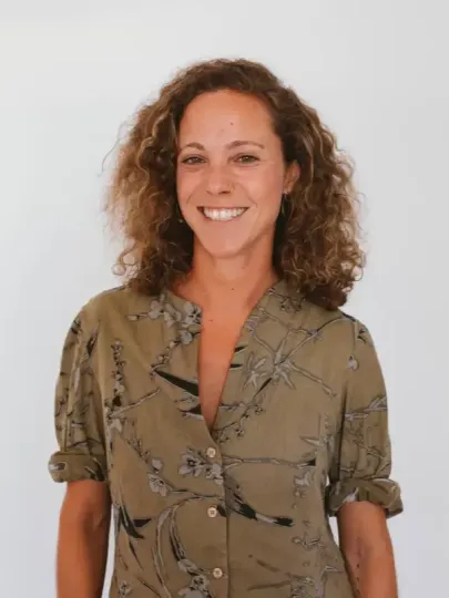 Una persona sonriente con cabello castaño rizado, que viste una camisa abotonada de color marrón claro con estampado