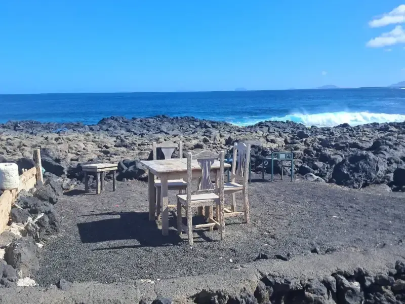 Una mesa y sillas de madera se asientan sobre un paisaje volcánico oscuro y rocoso, con vistas al océano azul