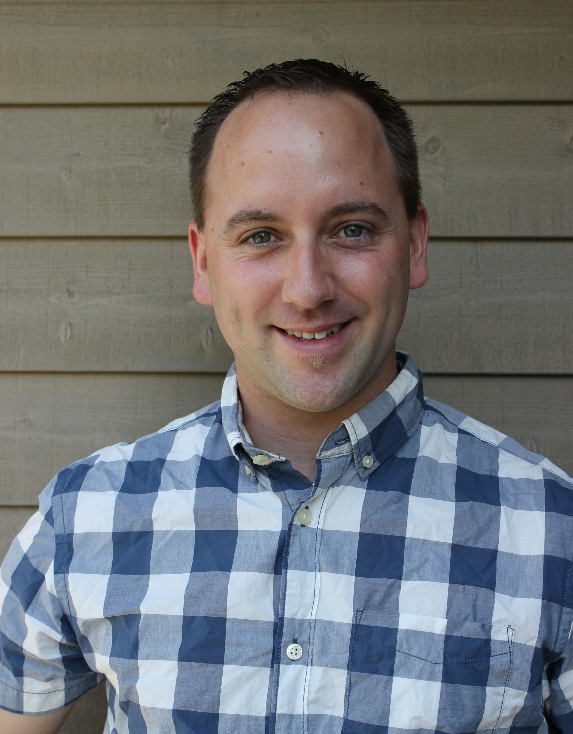Ein Mann in einem blau-weiß karierten Hemd lächelt vor einer holzgetäfelten Wand.
