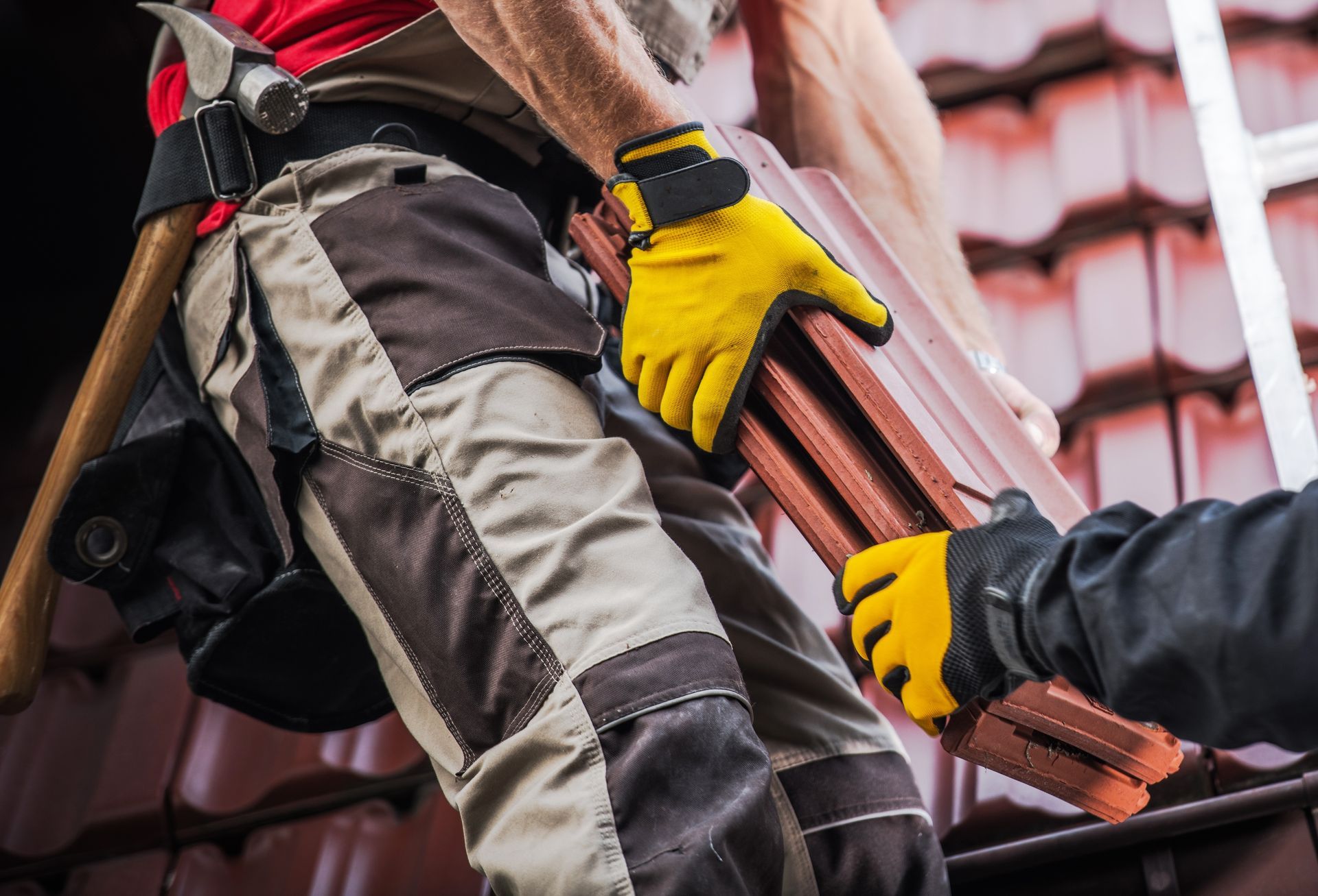 Des couvreurs en pantalon de travail et gants jaunes transportent des tuiles en céramique sur un chantier.