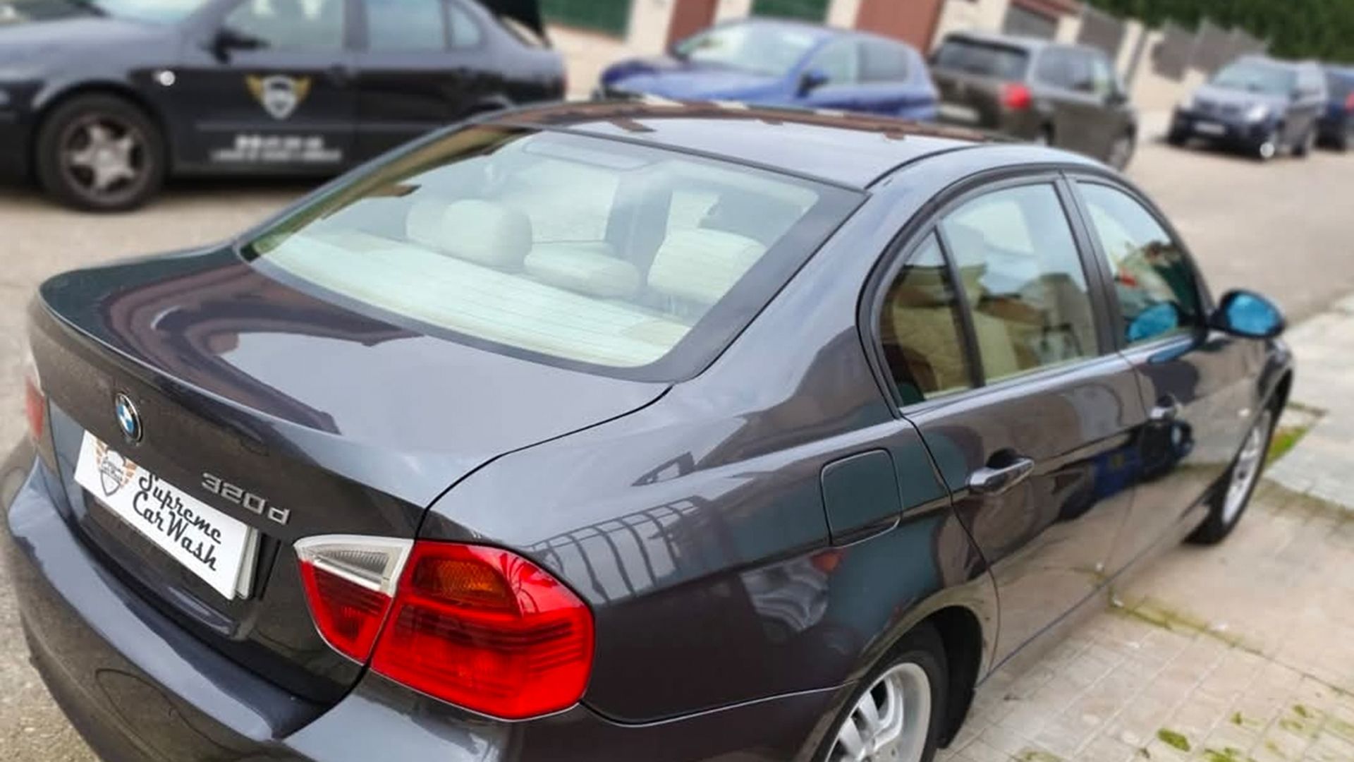 Un sedán BMW Serie 3 de color gris oscuro estacionado en una calle, con otros vehículos visibles al fondo.