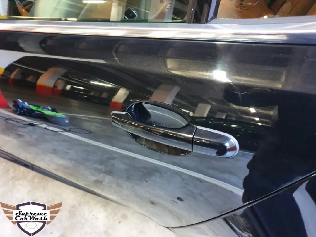 Primer plano de la puerta de un coche, negra y brillante, con tirador cromado, que refleja el aparcamiento cubierto circundante.