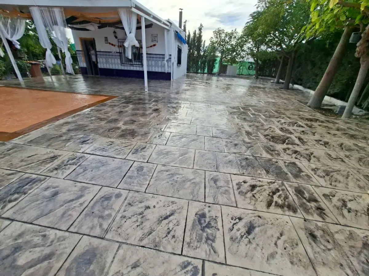 Toma exterior de un patio de hormigón húmedo y texturizado con un automóvil estacionado al fondo y césped verde a la derecha.