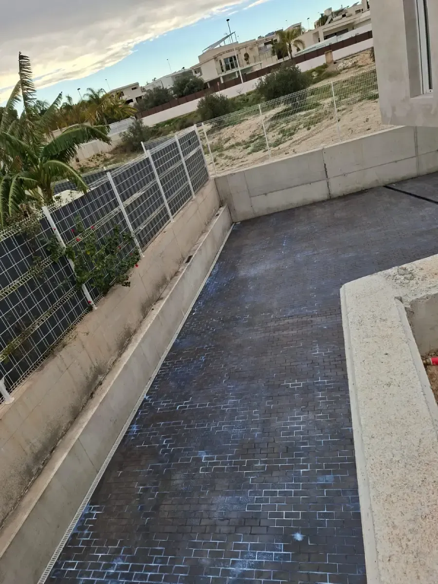 Un sendero oscuro con un diseño de ladrillos conduce al exterior, junto a un muro de contención y una valla de alambre con vegetación.