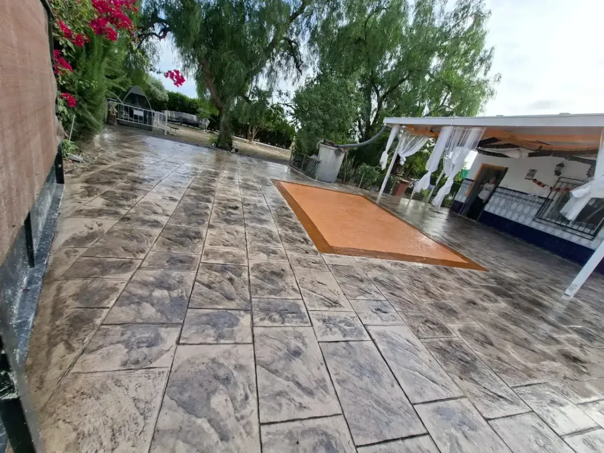 Patio de hormigón estampado con una zona rectangular de color naranja, que incluye un área de patio sombreada, árboles y vegetación a lo lejos.