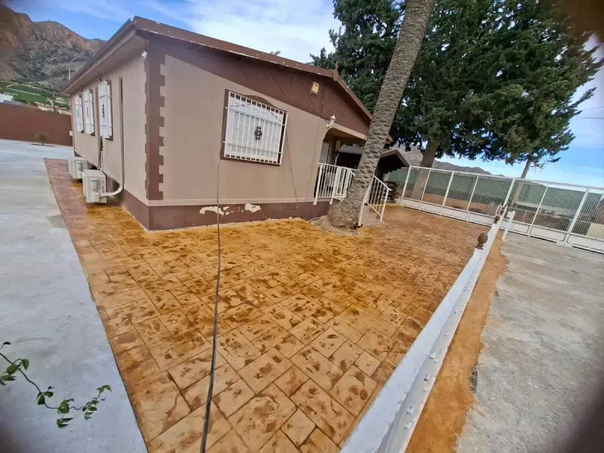 Camino de hormigón estampado con motivos cuadrados y color canela. Al fondo se ve un edificio y algo de tierra.