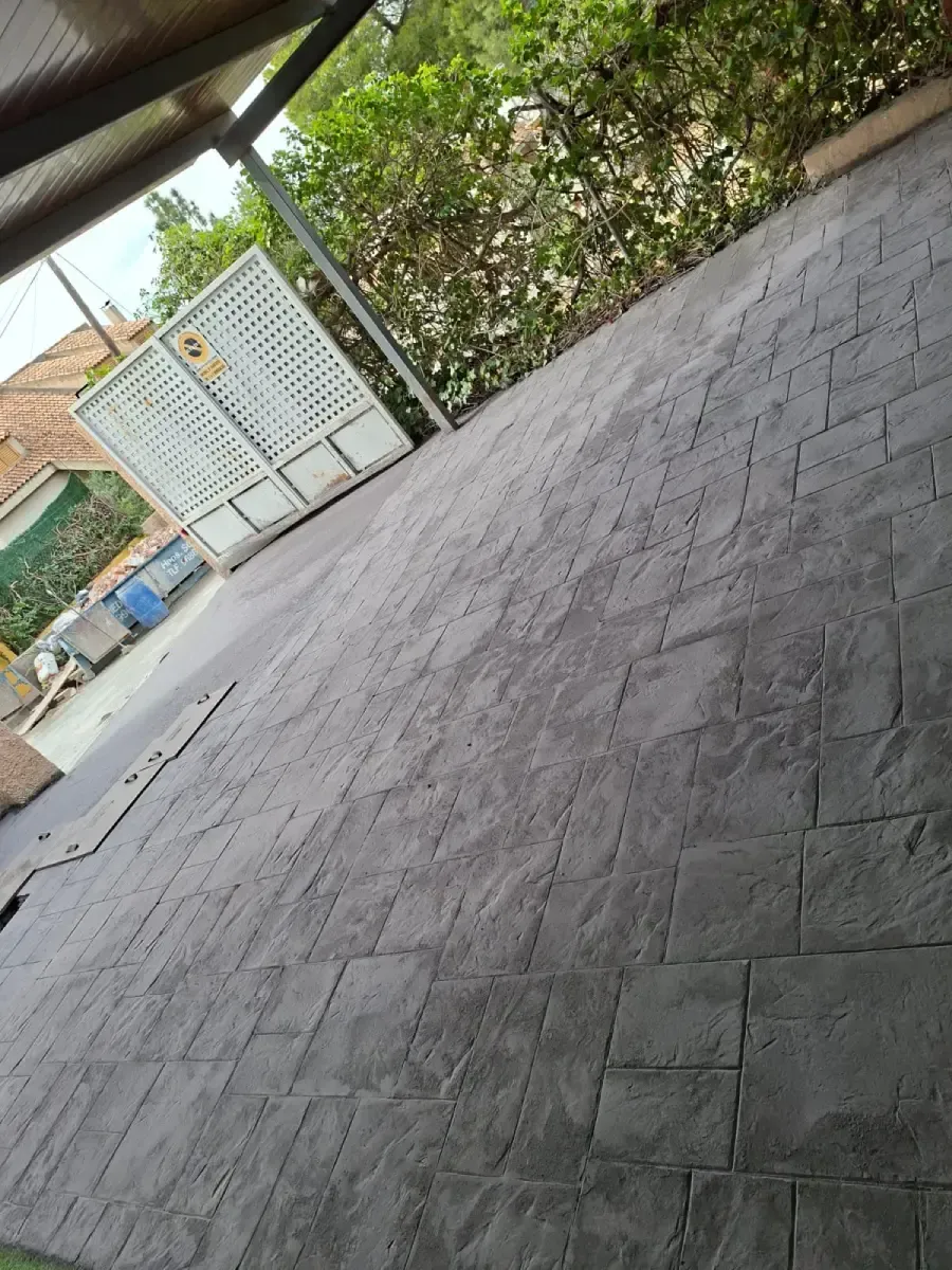 Primer plano de un suelo de hormigón estampado de color gris oscuro con un patrón cuadrado, situado en el exterior, cerca de una valla y un seto.