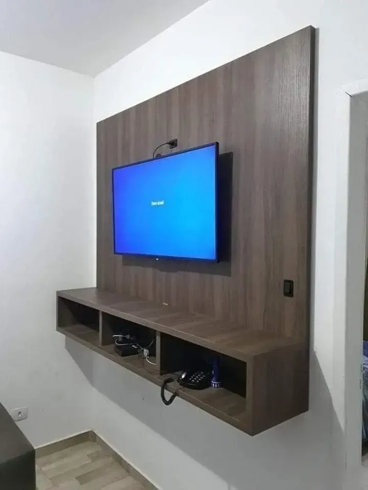 Televisor montado en un panel de pared de madera oscura con un estante flotante debajo; la pantalla es azul.