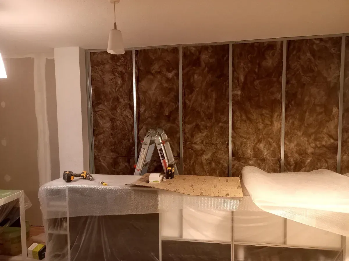 Pared interior en construcción con aislamiento entre montantes metálicos; herramientas sobre mostrador cubierto.