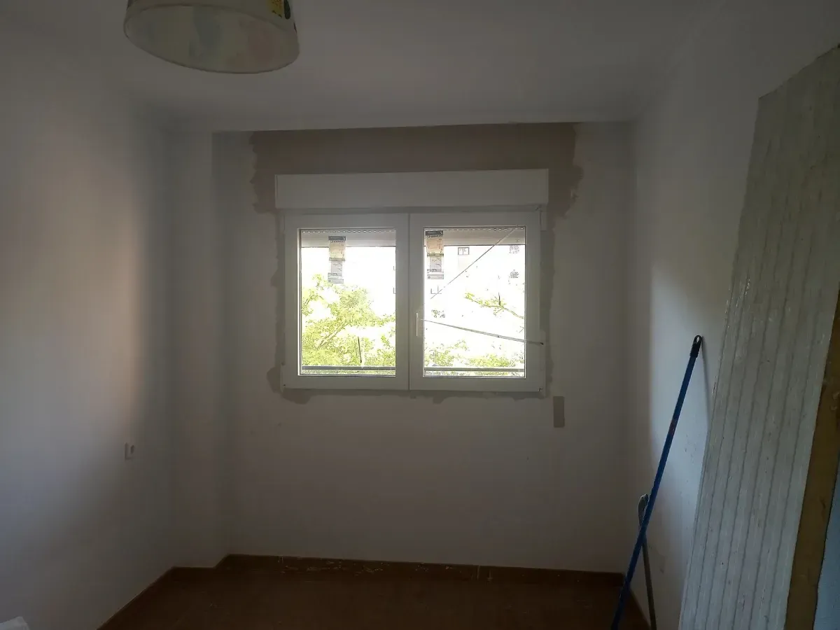 Habitación vacía con ventana; paredes blancas, suelo marrón y una escoba azul.