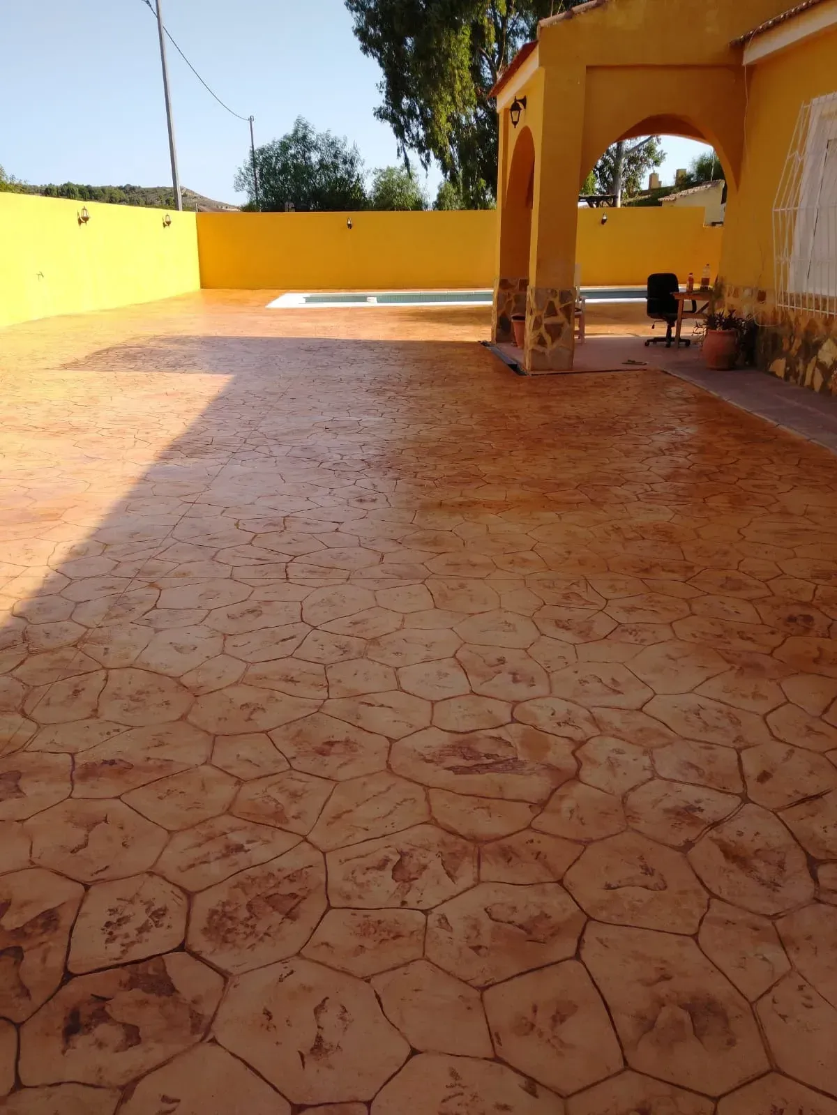 Patio de hormigón estampado de color naranja, edificio amarillo con entrada arqueada y una piscina al fondo.