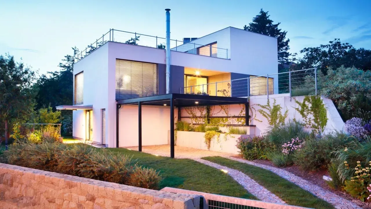 Casa blanca moderna con techo plano, entrada para vehículos y jardinería al anochecer.