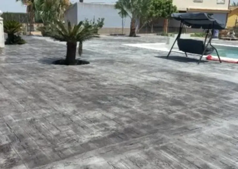 Patio de hormigón gris con árboles, un columpio y una piscina al fondo.