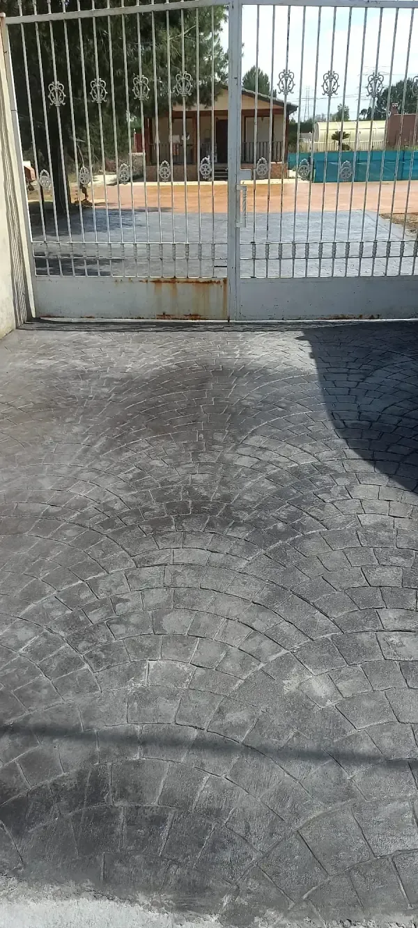 Entrada de cemento gris que conduce a una puerta metálica. Detrás de la puerta hay un edificio y algunos árboles.