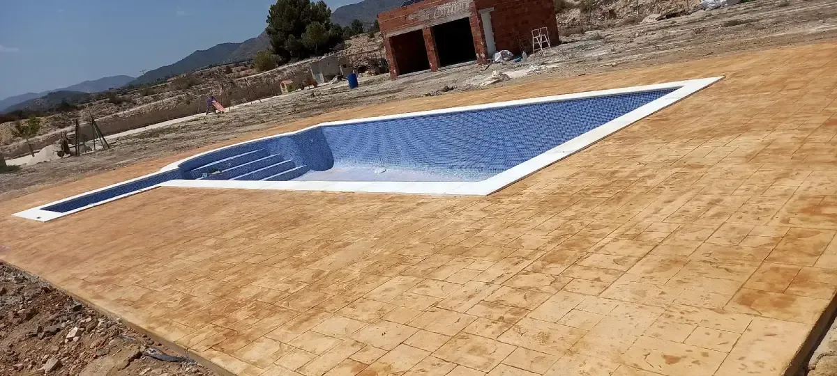 Una nueva piscina geométrica, con azulejos azules, se encuentra en una zona de arena cerca de un edificio de ladrillos