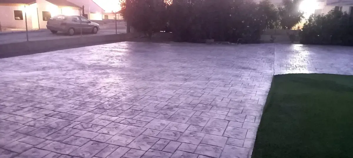 Toma exterior de un patio de hormigón húmedo y texturizado con un automóvil estacionado al fondo y césped verde a la derecha.