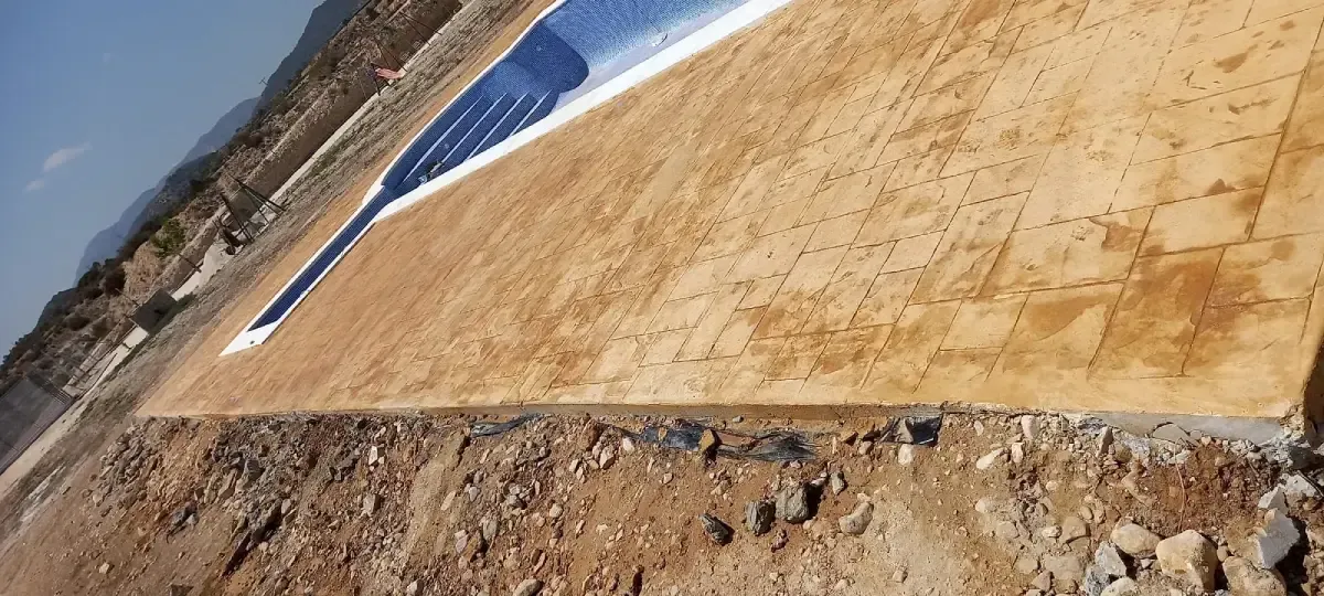 Una piscina parcialmente construida. Está rodeada de hormigón color canela, con montañas y cielo azul de fondo.
