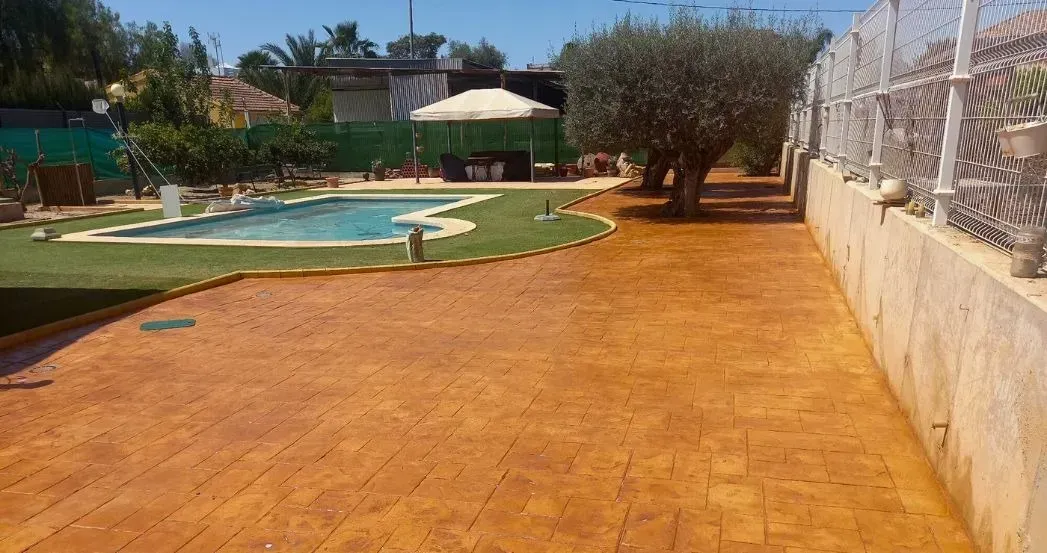 Camino de hormigón estampado con apariencia de ladrillo en un patio con piscina, cenador, árboles y cerca decorativa.