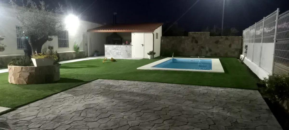 Patio trasero de noche con piscina, césped artificial y una pequeña construcción con techo de tejas.