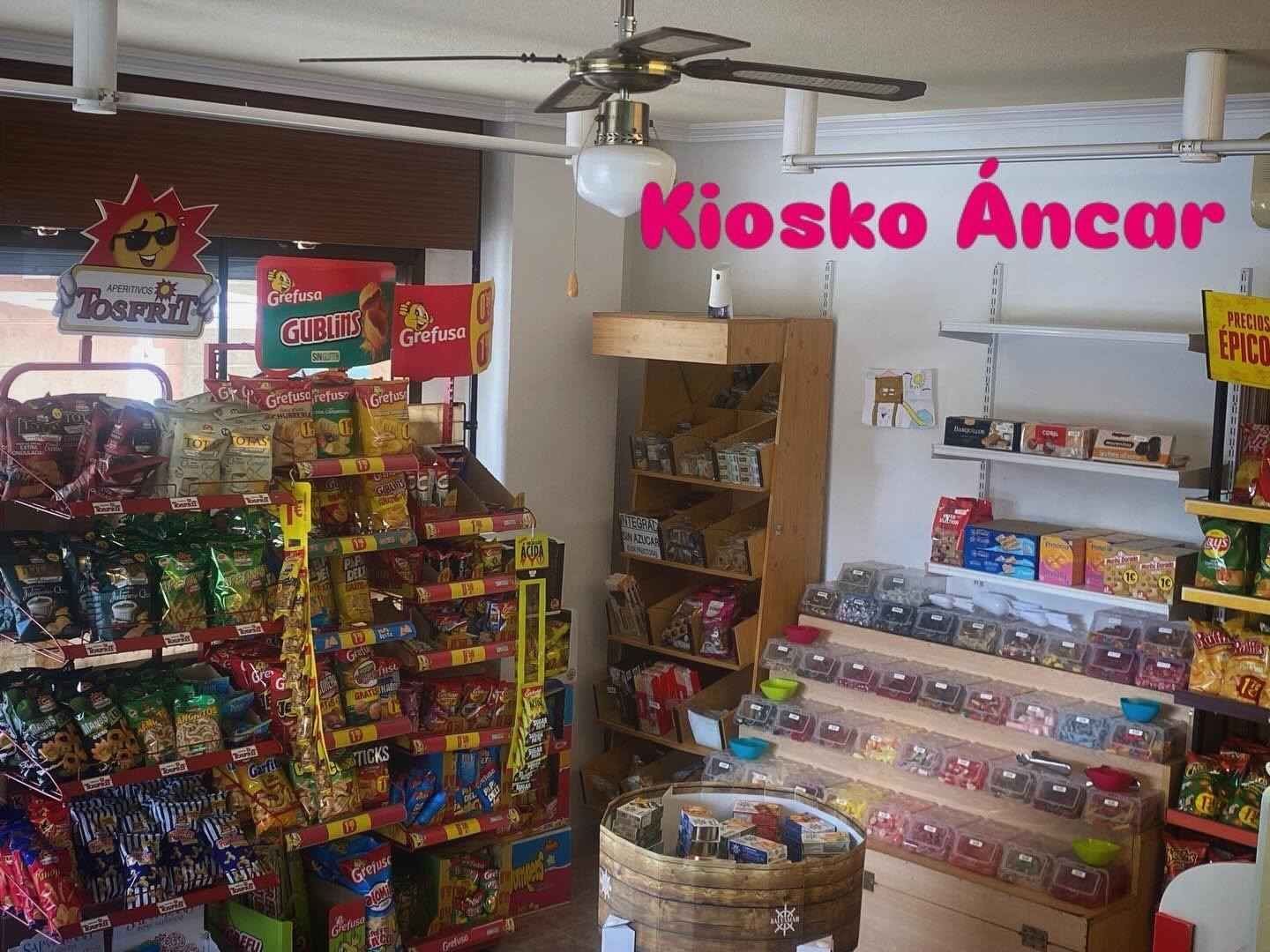 Quiosco de aperitivos colorido con estantes de caramelos y patatas fritas, etiquetado como “Kiosko Ancar” en el interior de una pequeña tienda.