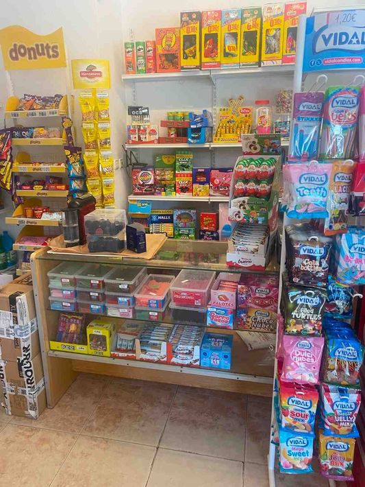 Pequeño escaparate de tienda de conveniencia con aperitivos, caramelos y golosinas envasadas de colores en estantes y percheros.