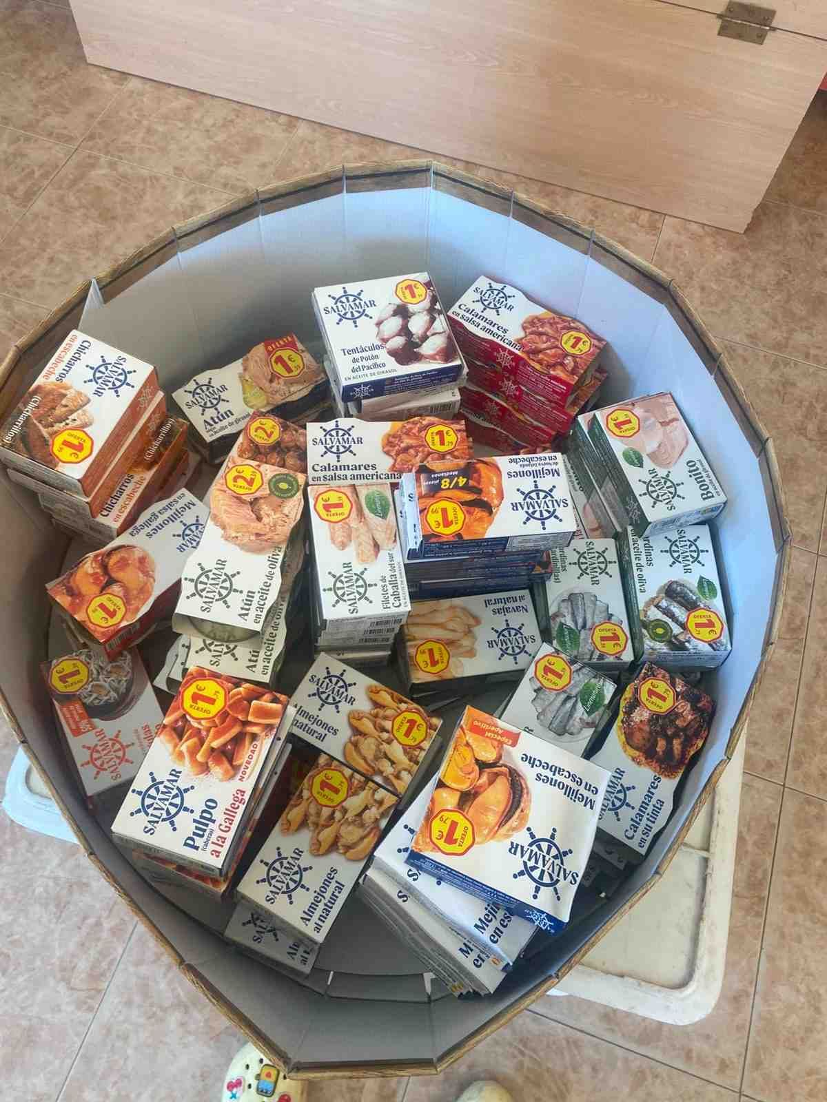 Caja de cartón abierta llena de paquetes surtidos de aperitivos o dulces de colores sobre un suelo de baldosas.
