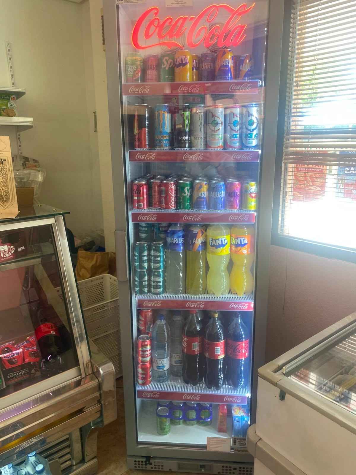 Vitrina refrigerada de Coca-Cola con coloridas latas y botellas de refresco en una pequeña tienda.