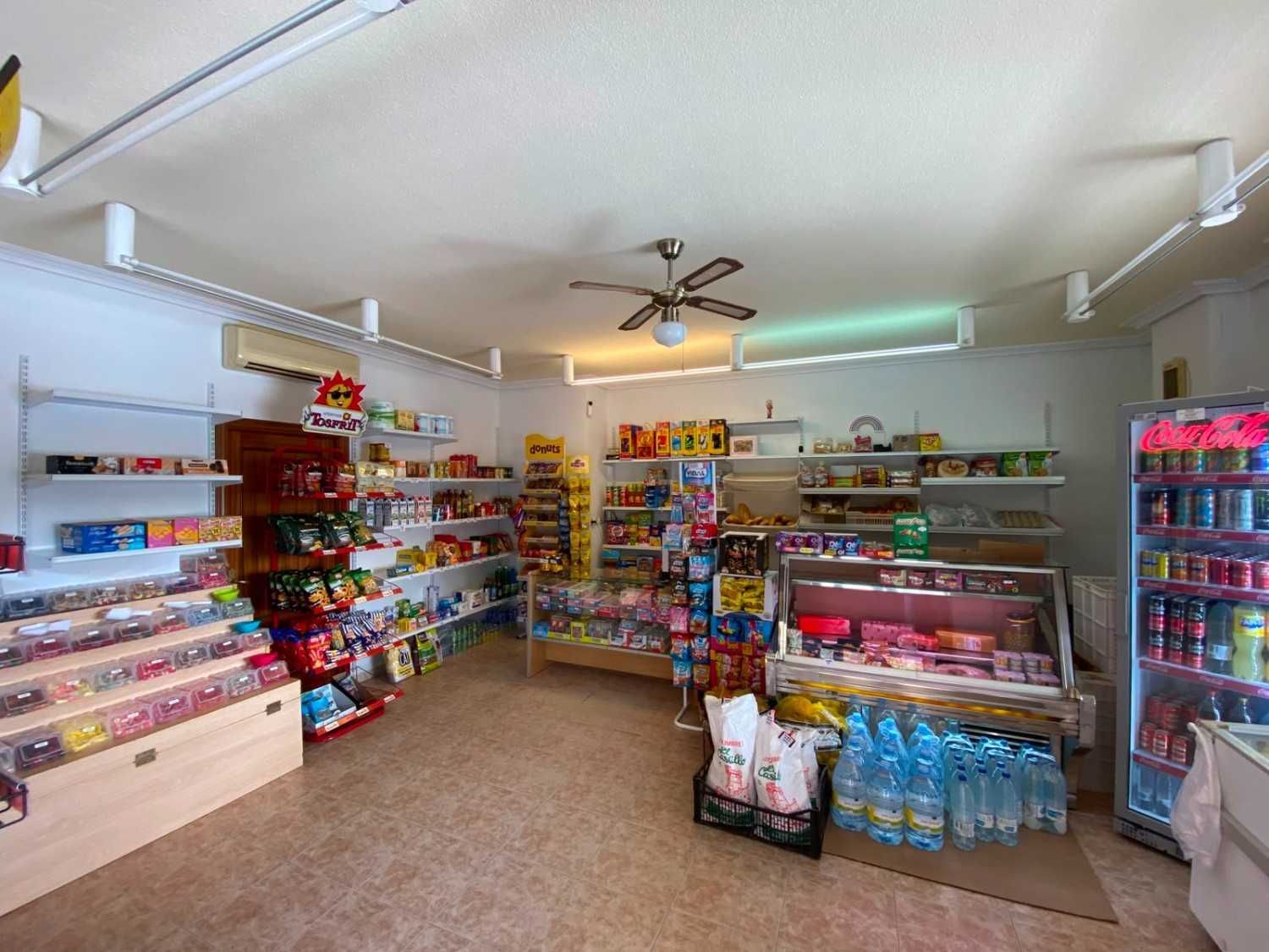 Pasillo de tienda de conveniencia con estantes de bocadillos, dulces, bebidas y artículos para el hogar bajo luces brillantes.