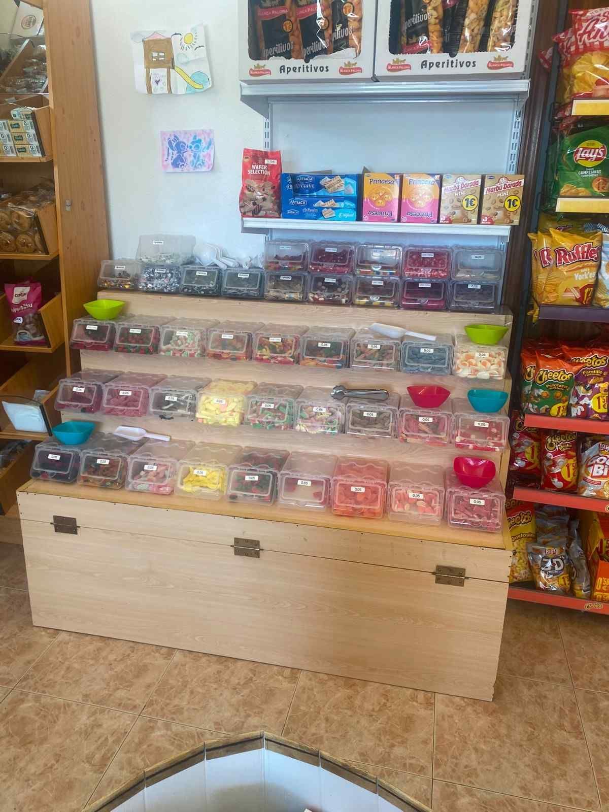 Estantería pequeña de una tienda con artículos empaquetados de colores y filas de recipientes transparentes para dulces debajo de las fotos.