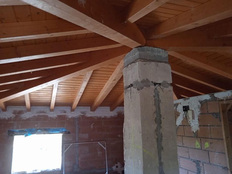 Ático sin terminar, con vigas de madera a la vista, paredes de ladrillo y una columna de hormigón, iluminado por una ventana luminosa.