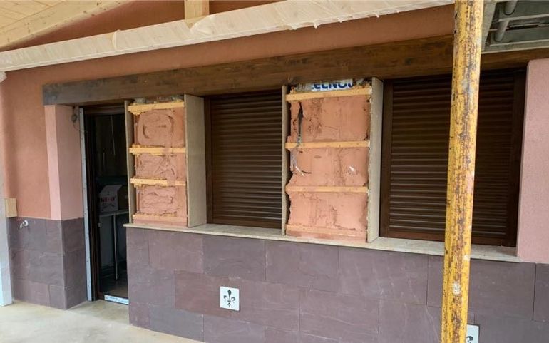 Ventana tapiada en un edificio con contraventanas marrones y una fachada manchada, vista detrás de una columna