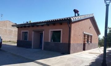 Pequeña casa de estuco color melocotón, con tejado de tejas y una persona de pie sobre él bajo un sol radiante