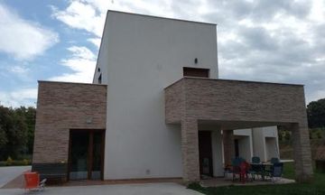 Casa moderna con una sección central blanca y alas de ladrillo, vista de frente, con un patio y sillas de exterior.