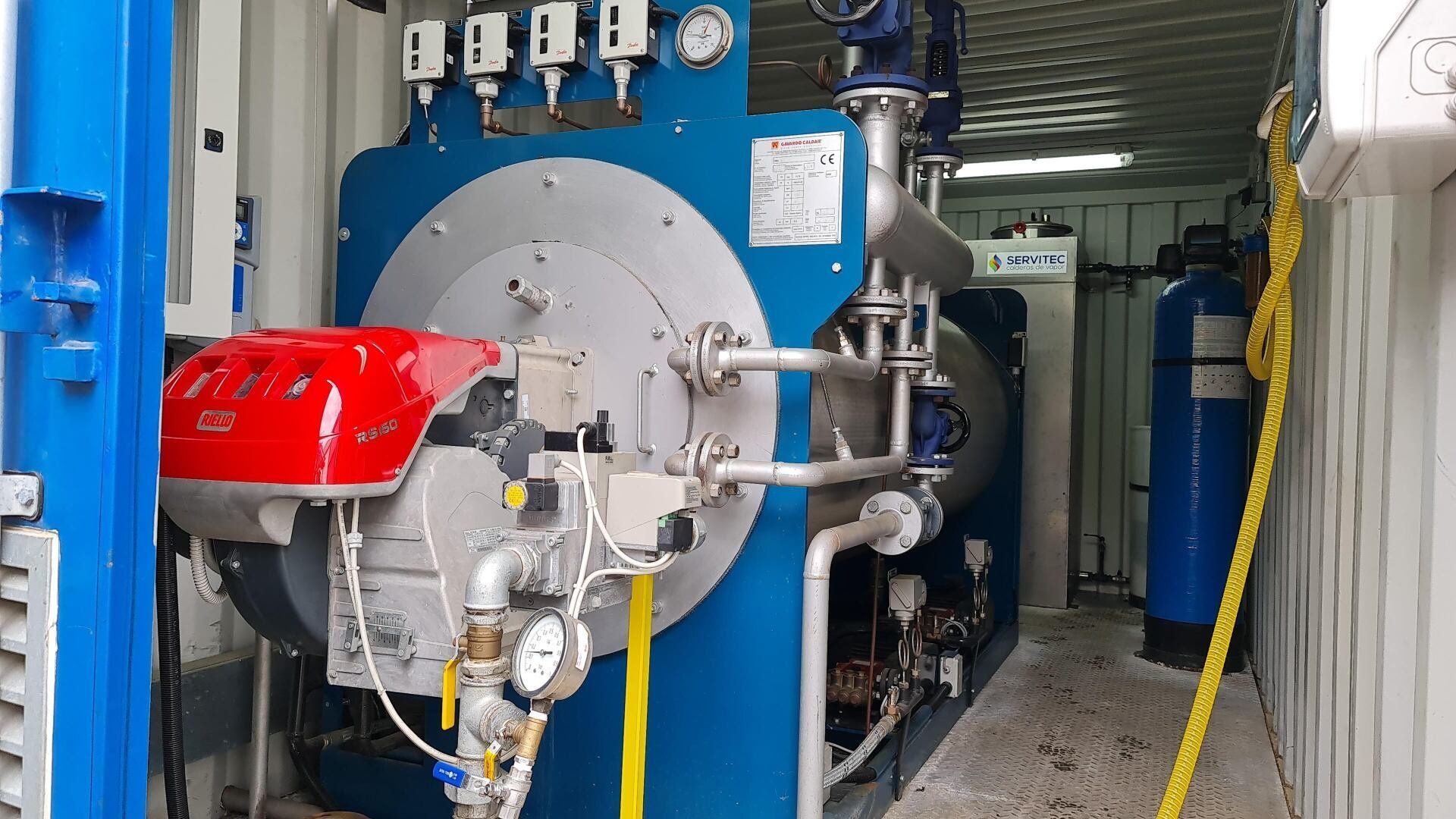 Una gran caldera industrial azul con un quemador rojo dentro de un contenedor blanco. Se ven las tuberías y los medidores.