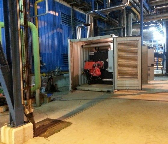 Una gran instalación industrial muestra un contenedor abierto que alberga quemadores y tuberías rojas. Al fondo, se ven tuberías azules y verdes.