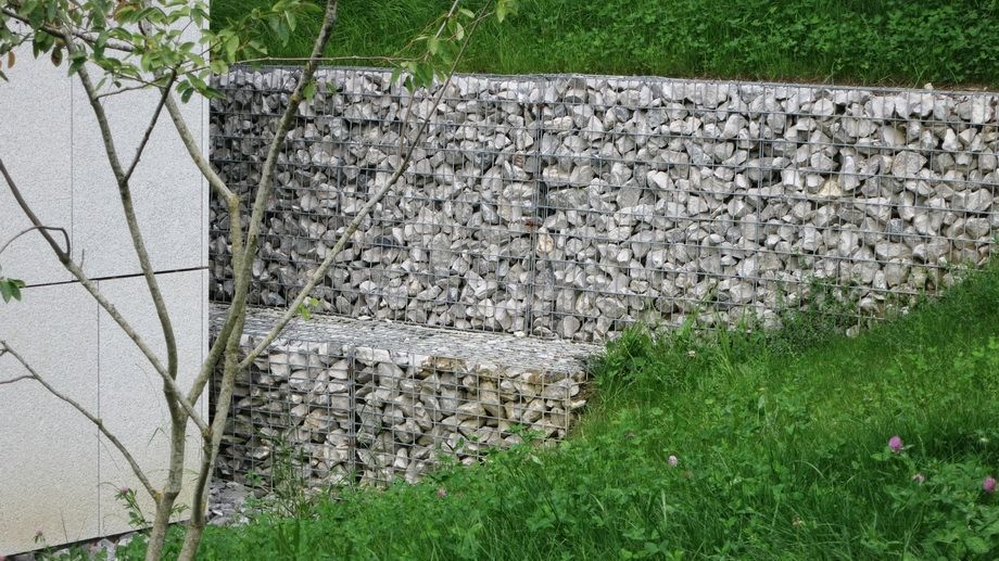 Mur en gabions de pierre jouxtant une structure en béton, avec de l'herbe verte.