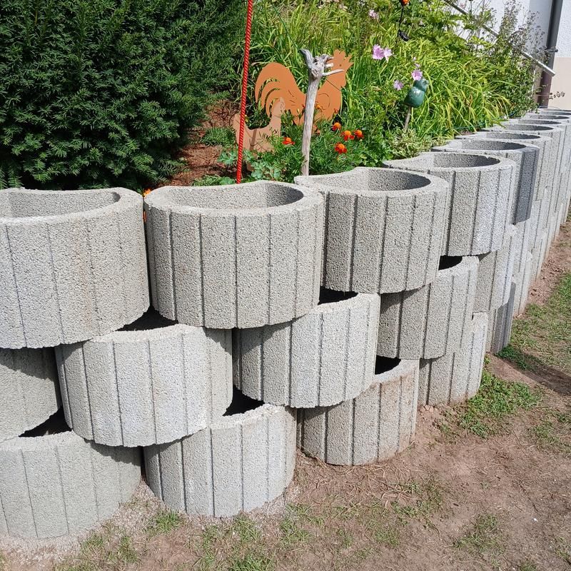 Des blocs de béton empilés forment un mur de soutènement de jardin, avec des plantes en arrière-plan.