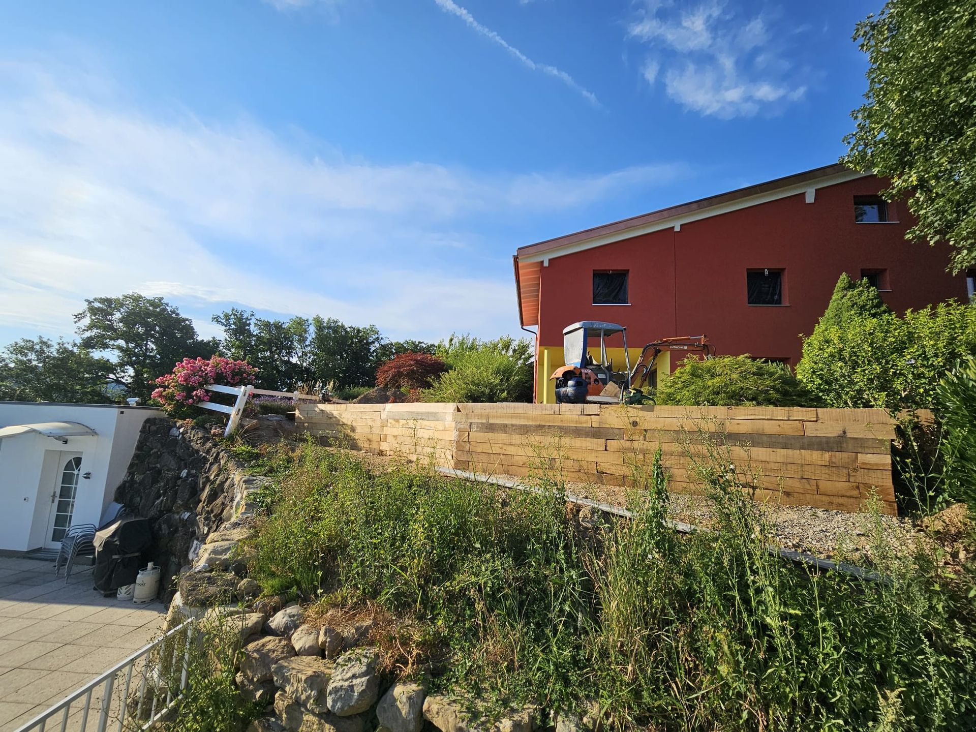 Maison rouge à flanc de colline, entourée de murs de soutènement et aménagée. Ciel bleu.