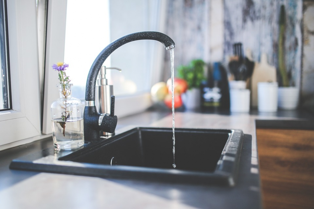 Fregadero de cocina negro con agua que fluye de un grifo curvo; ventana con jarrón y flores a la izquierda.